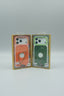 Two packaged phone cases displayed side by side in clear retail packaging. The left case is orange with a white camera cutout area and a thin metallic wire-like accent, while the right case is green with a similar white camera area and a thin multicolored wire accent.