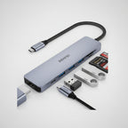 A silver and black USB-C hub with multiple ports, shown with various accessories including a USB-C cable, SD card, and USB flash drive on a white background.