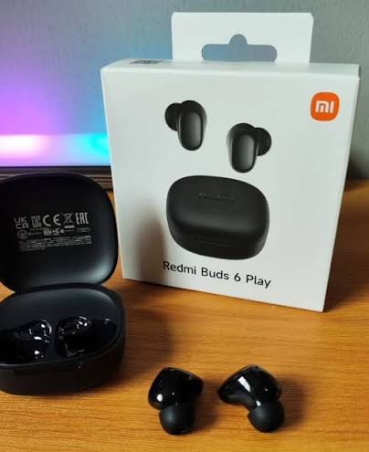 Black Xiaomi Redmi Buds 6 Play wireless earbuds shown with their open charging case, alongside the original white product packaging box on a wooden surface.