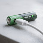 A green cylindrical rechargeable battery with a white USB charging cable plugged into it, resting on a light gray textured surface.