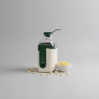 A small popcorn machine with a cream-colored body and green accents sits on a light countertop, with scattered popcorn kernels around it and a small bowl of popcorn nearby in a bright kitchen setting.