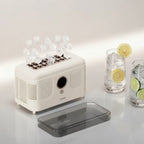 A compact white ice maker machine with a transparent lid placed in front, shown on a clean light background. Several clear ice cubes are visible above the top tray, and two glass drinks with lemon and lime slices sit to the right as styling props.