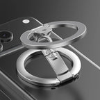 A metallic silver phone grip ring with a built-in kickstand, shown attached to the back of a black smartphone on a dark reflective surface.